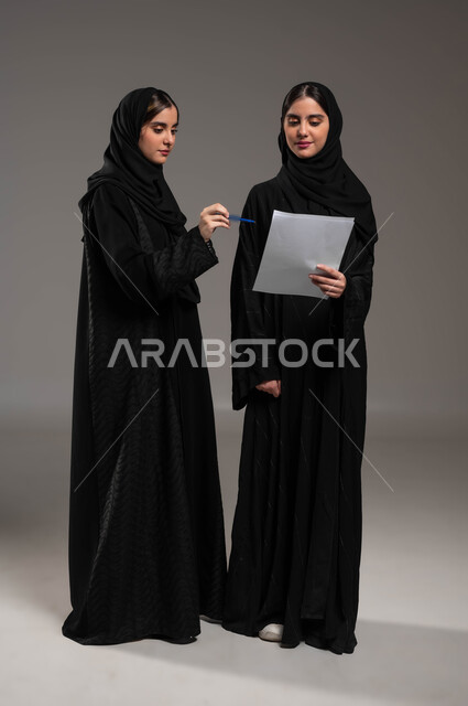 الوحده والعمل الجماعي والمشاركة، اجتماع زملاء العمل في مقر عملهم، بورتريه لتوءم بنات عربيتان خليجيتان سعوديتان ترتديان الحجاب والعباءة السوداء تقومان بمناقشة أمور العمل، الإشارة بالإصبع على أوراق المشاريع، صورة الجسم بالطول الكامل، خلفية رمادية