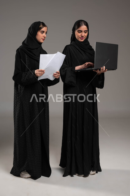 المساعدة في الأعمال ومناقشتها عبر الاجهزة المتطورة، بورتريه لتوءم بنات عربيتان خليجيتان سعوديتان ترتديان الحجاب والعباءة السوداء احدهما تمسك أوراق العمل والأخرى تحمل الحاسب المحمول, اجتماع زملاء عمل، صورة الجسم بالطول الكامل، خلفية رمادية