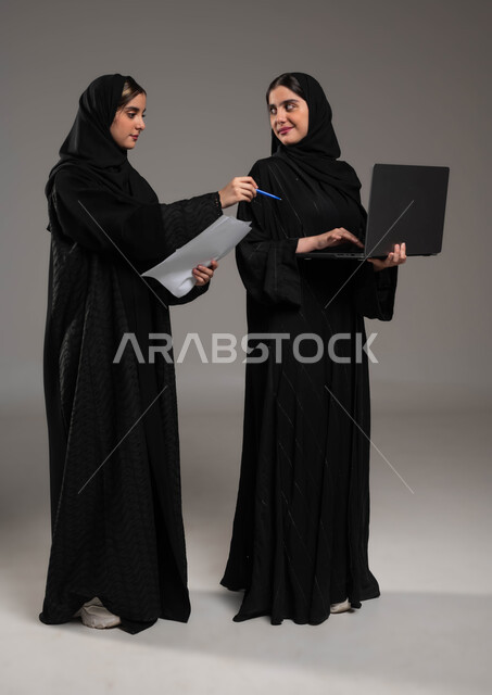 مناقشة الاعمال عبر الاونلاين, بورتريه لبنتان توءم عربيتان سعوديتان خليجيتان ترتديان الحجاب والعباءة احداهما تمسك الحاسب المحمول والاخرى اوراق العمل, الاشارة بالاصبع على الحاسوب، صورة الجسم بالطول الكامل, خلفية رمادية