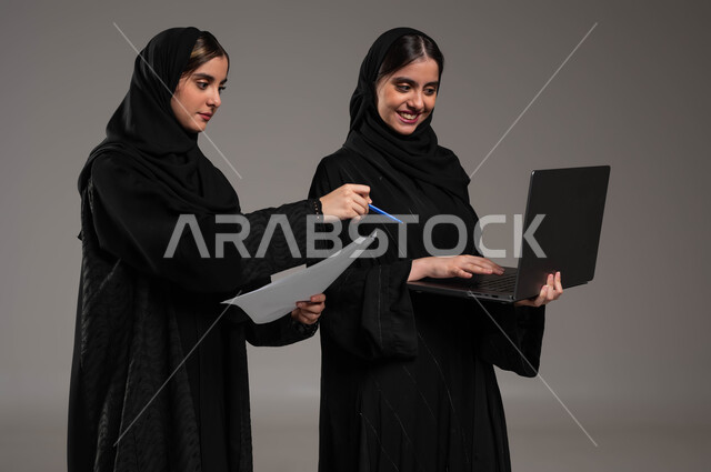مناقشة الاعمال عبر الاجهزة المتطورة, محاولة إيجاد فكرة ما عبر الأونلاين, الاشارة بالاصبع على الحاسوب, بورتريه مقرب لبنتان عربيتان سعوديتان خليجيتان ترتديان الحجاب والعباءة احداهما تمسك الحاسب المحمول والاخرى اوراق العمل, خلفية رمادية
