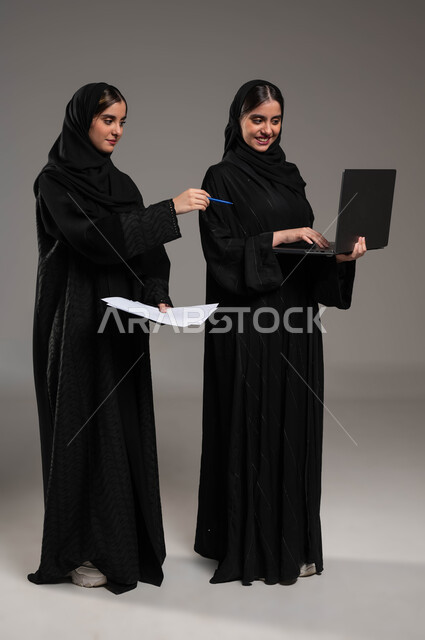 محاولة إيجاد فكرة ما عبر الأونلاين, الاشارة بالاصبع على الحاسوب, مناقشة الاعمال عبر الاجهزة المتطورة, بورتريه لبنتان عربيتان سعوديتان خليجيتان ترتديان الحجاب والعباءة احداهما تمسك الحاسب المحمول والاخرى اوراق العمل, صورة الجسم بالطول الكامل, خلفية رمادية
