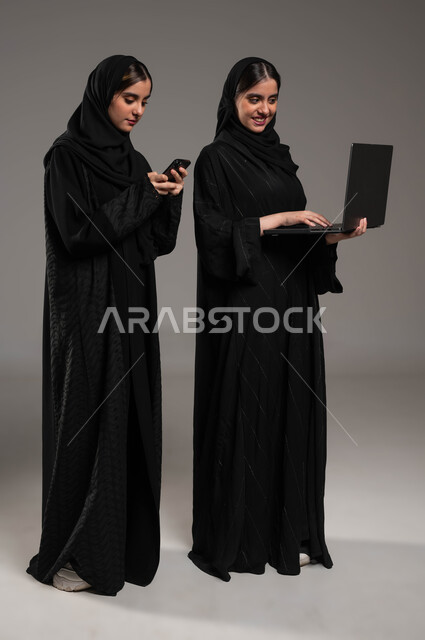 التواصل التكنولوجي الحديث عبر السوشال ميديا, استخدام الأجهزة الالكترونية الحديثة, بورتريه مقرب لبنتان عربيتان سعوديتان خليجيتان ترتديان الحجاب والعباءة احداهما تمسك الابتوب والاخرى الجوال, صورة الجسم بالطول الكامل, خلفية رمادية