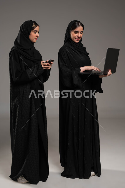 التواصل مع الأصدقاء عبر السوشال ميديا, استخدام الحاسوب في تطوير العمل, دور التقنيات الحديثة في ايجاد المعلومات, بورتريه لبنتان عربيتان سعوديتان خليجيتان ترتديان الحجاب والعباءة احداهما تمسك الابتوب والاخرى الهاتف المحمول, صورة الجسم بالطول الكامل, خلفية رمادية