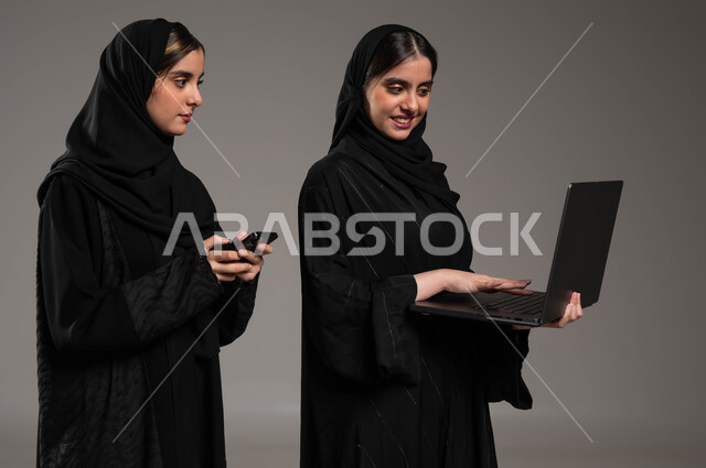 The use of computers in developing work, the role of modern technologies in finding information, a close-up portrait of two Saudi Arabian Gulf girls wearing the hijab and abaya, one holding a laptop and the other a mobile phone, communicating with friends via social media, gray background