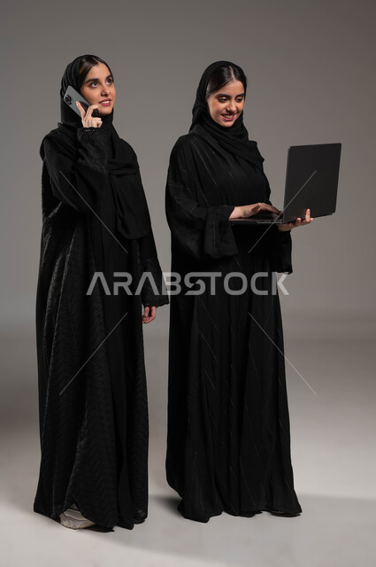 اجراء مكالمة هاتفية عبر الجوال, استخدام التقنيات الحديثة للتواصل الاجتماعي, بورتريه لبنتان توءم سعوديتان خليجيتان عربيتان ترتديان الحجاب والعباءة احداهما تمسك الابتوب والاخرى الهاتف المحمول, صورة الجسم بالطول الكامل, خلفية رمادية