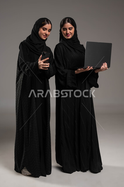 التواصل مع الأصدقاء عبر السوشال ميديا, استخدام الأجهزة والتقنيات الحديثة, بورتريه لبنتان توءم سعوديتان خليجيتان عربيتان ترتديان الحجاب والعباءة احداهما تمسك الحاسب المحمول والاخرى الهاتف, إجراء مكالمة فيديو عبر الجوال, صورة الجسم بالطول الكامل, خلفية رمادية