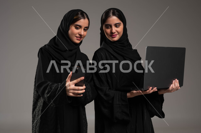 استخدام الأجهزة والتقنيات الحديثة, بورتريه مقرب لبنتان توءم سعوديتان خليجيتان عربيتان ترتديان الحجاب والعباءة احداهما تمسك الحاسب المحمول والاخرى الهاتف المحمول, إجراء مكالمة فيديو عبر الجوال, التواصل مع الأصدقاء عبر السوشال ميديا, خلفية رمادية