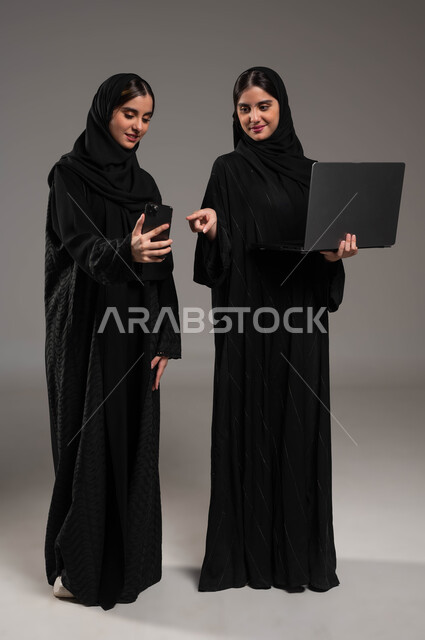 استخدام الاجهزة المتطورة والتقنيات الحديثة, بورتريه لبنتان عربيتان سعوديتان خليجيتان ترتديان الحجاب والعباءة احدهما تمسك بيدها الحاسب المحمول والاخرى الهاتف, إجراء مكالمة فيديو والاشارة بالاصبع الى الجوال, صورة الجسم بالطول الكامل, خلفية رمادية