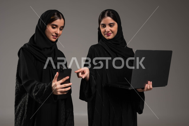 إجراء مكالمة فيديو والاشارة بالاصبع الى الجوال, مشاهدة محتويات مفيدة, استخدام الاجهزة المتطورة والتقنيات الحديثة, بورتريه مقرب لبنتان عربيتان سعوديتان خليجيتان ترتديان الحجاب والعباءة احدهما تمسك بيدها الحاسب المحمول والاخرى الهاتف, خلفية رمادية