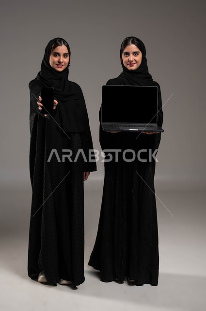 استخدام الأجهزة التقنية المتطورة, ايماءات الفرح والسعادة, دور التقنيات الحديثة في عملية البحث عن المعلومات المفيدة, بورتريه لبنتان توءم عربيتان سعوديتان خليجيتان ترتديان الحجاب والعباءة احداهما تمسك بيدها الحاسب المحمول والاخرى الهاتف الجوال بشاشة سوداء فارغة, صورة الجسم بالطول الكامل, خلفية رمادية