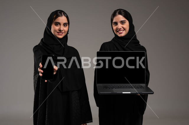 ايماءات الفرح والسعادة, دور التقنيات الحديثة في عملية البحث عن المعلومات المفيدة, استخدام الأجهزة التقنية المتطورة, بورتريه مقرب لبنتان توءم عربيتان سعوديتان خليجيتان ترتديان الحجاب والعباءة احداهما تمسك بيدها الحاسب المحمول والاخرى الهاتف الجوال بشاشة سوداء فارغة, خلفية رمادية