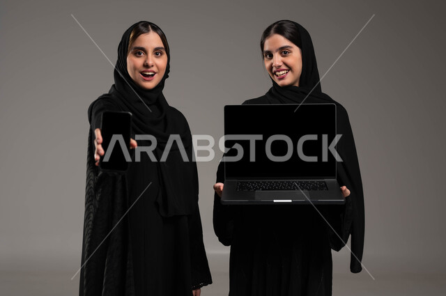 دور التقنيات الحديثة في عملية البحث عن المعلومات المفيدة, استخدام الأجهزة التقنية المتطورة, بورتريه مقرب لبنتان توءم عربيتان سعوديتان خليجيتان ترتديان الحجاب والعباءة احداهما تمسك بيدها الحاسب المحمول والاخرى الهاتف الجوال بشاشة سوداء فارغة, ايماءات الفرح والحماس, خلفية رمادية
