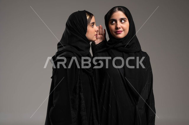 ايماءات الانصات والتركيز بالكلام، النميمة والتحدث عن شخص ما، التحدث بصوت منخفض، بورتريه مقرب لشابة عربية خليجية سعودية ترتدي الحجاب والعباءة تهمس بإذن أختها التوأم، مفهوم الكتمان وحفظ الأسرار بين الشقيقات، خلفية رمادية