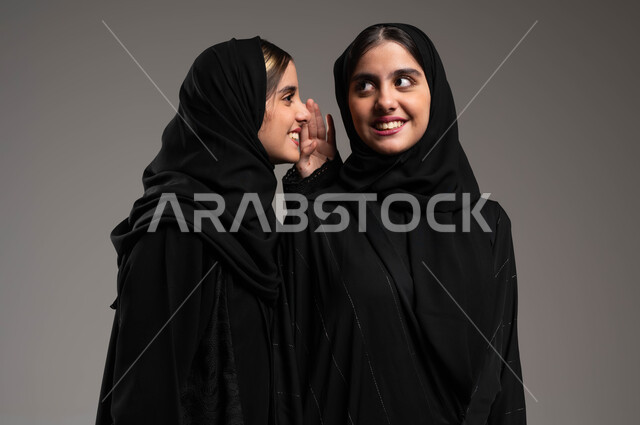 النميمة والتحدث عن شخص ما، ايماءات الانصات والتركيز بالكلام، التحدث بصوت منخفض، بورتريه مقرب لشابة عربية خليجية سعودية ترتدي الحجاب والعباءة تهمس بإذن أختها التوأم، مفهوم الكتمان وحفظ الأسرار بين الشقيقات، خلفية رمادية