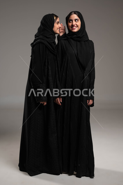 النميمة والتحدث عن شخص ما، التحدث بصوت منخفض، بورتريه لشابة عربية خليجية سعودية ترتدي الحجاب والعباءة تهمس بإذن أختها التوأم، مفهوم الكتمان وحفظ الأسرار بين الشقيقات،  ايماءات الانصات والتركيز بالكلام، صورة الجسم بالكامل، خلفية رمادية