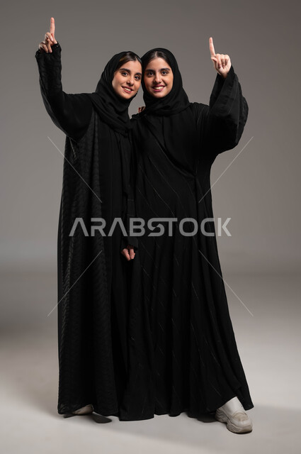 الوقوف باستقامة وثقة بالنفس، الاشارة بأصبع السبابه نحو الأعلى بإيماءات السرور، الاناقة والاهتمام بالمظهر الخارجي، بورتريه لأختين توءم عربيتين خليجيتين سعوديتين محجبتين مبتسمتين ترتديان العباءة السوداء تنظران الى الكاميرا، صورة لكامل الجسم، خلفية رمادية