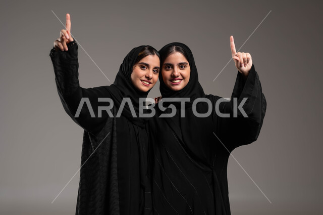 الاشارة بأصبع السبابه نحو الأعلى بإيماءات السرور، الوقوف باستقامة وثقة بالنفس، الاناقة والاهتمام بالمظهر الخارجي، بورتريه مقرب لأختين توءم عربيتين خليجيتين سعوديتين محجبتين مبتسمتين ترتديان العباءة السوداء تنظران الى الكاميرا، خلفية رمادية
