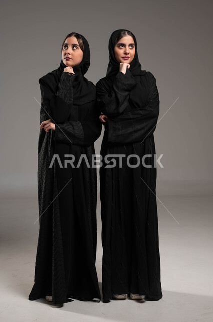 Absent-mindedness and thinking, trying to find a solution to a problem, feeling and feeling bored and fed up, waiting for something, a portrait of two Saudi Gulf Arab twin sisters, veiled, wearing the black abaya, looking up with gestures of boredom and thought, full-body portrait, gray background