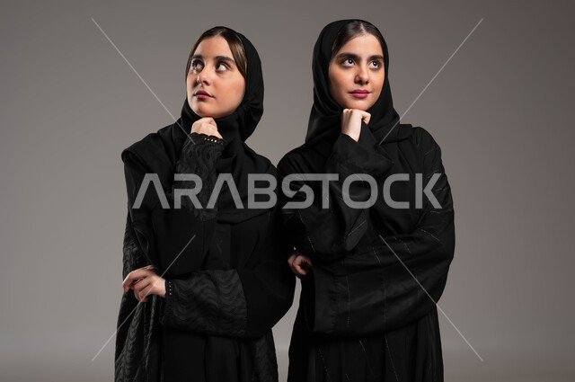 محاولة إيجاد حل لمشكلة ما، الاحساس والشعور بالضجر والزهق، انتظار شيء ما، بورتريه مقرب لاختين توءم عربيتين خليجيتين سعوديتين محجتبتين ترتديان العباءة السوداء تنظران للأعلى بإيماءات الملل والتفكير، شرود الذهن والتفكير، خلفية رمادية