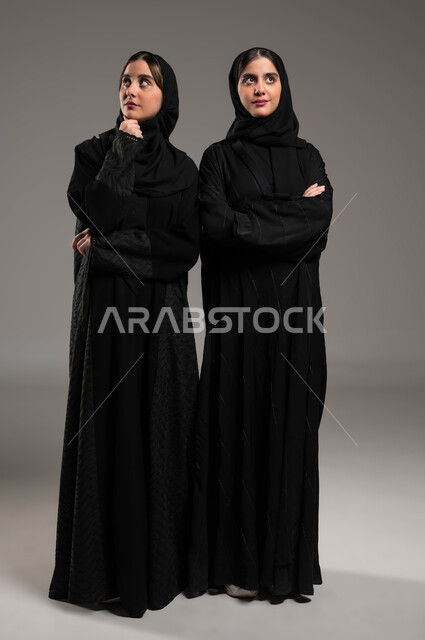 شرود الذهن والتفكير، محاولة إيجاد حل لمشكلة ما، الاحساس والشعور بالضجر والزهق، انتظار شيء ما، بورتريه لاختين توءم عربيتين خليجيتين سعوديتين محجتبتين ترتديان العباءة السوداء تنظران للأعلى بإيماءات الملل والتفكير، صورة لكامل الجسم، خلفية رمادية