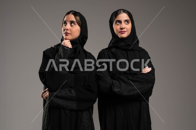 Trying to find a solution to a problem, feeling and feeling bored and fed up, waiting for something, a close-up portrait of two veiled Saudi Gulf Arab twin sisters, wearing the black abaya, looking up with gestures of boredom and thought, absent-mindedness and thinking, gray background