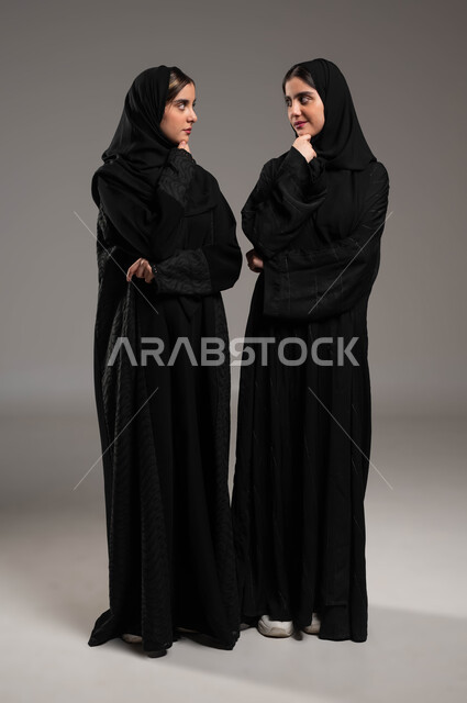 انتظار شيء ما، شرود الذهن والتفكير، محاولة إيجاد حل لمشكلة ما، الاحساس والشعور بالملل، بورتريه لاختين توءم عربيتين خليجيتين سعوديتين محجتبتين ترتديان العباءة السوداء تنظران لبعضهما بإيماءات التفكير، صورة لكامل الجسم، خلفية رمادية