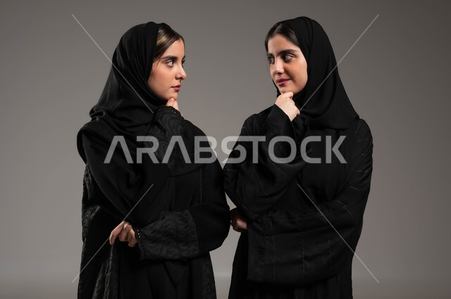 Waiting for something, absent-mindedness and thinking, trying to find a solution to a problem, feeling and feeling bored, a close-up portrait of two Saudi Gulf Arab twin sisters, veiled, wearing the black abaya, looking at each other with thinking gestures, gray background.