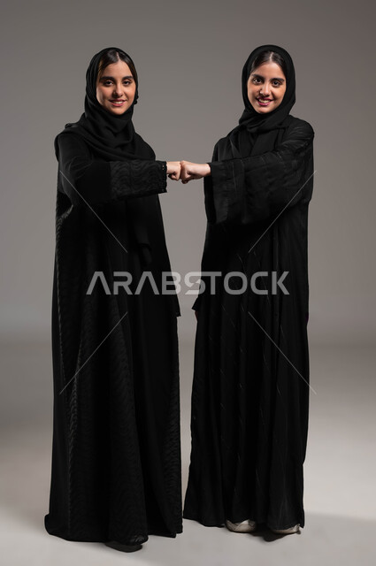 الحفاظ على المسافات بين الاشخاص، اتباع احترازات السلامة، بورتريه لشابتين توءم عربيتين خليجيتين سعودتين محجبتين تقومان بالتصافح بقبضة اليد، الالتزام بإجراءات الوقاية من فايروس كورونا، النظر إلى الكاميرا بابتسامة، صورة لكامل الجسم، خلفية رمادية