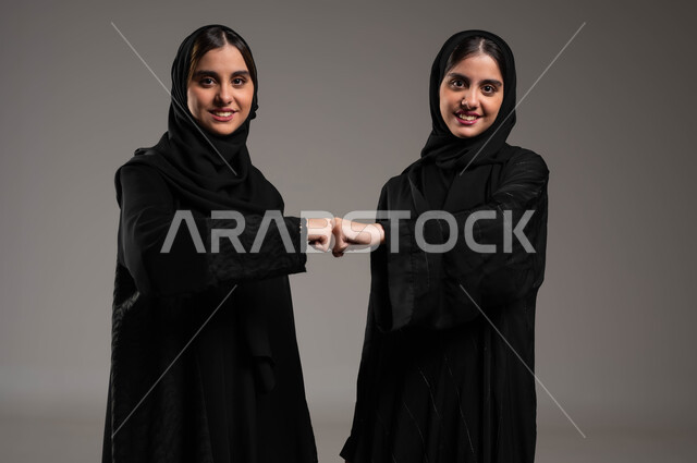 التباعد واتباع احترازات السلامة، الحفاظ على المسافات بين الاشخاص، بورتريه لشابتين توءم عربيتين خليجيتين سعودتين محجبتين تقومان بالتصافح بقبضة اليد، الالتزام بإجراءات الوقاية من فايروس كورونا، النظر إلى الكاميرا بابتسامة، خلفية رمادية