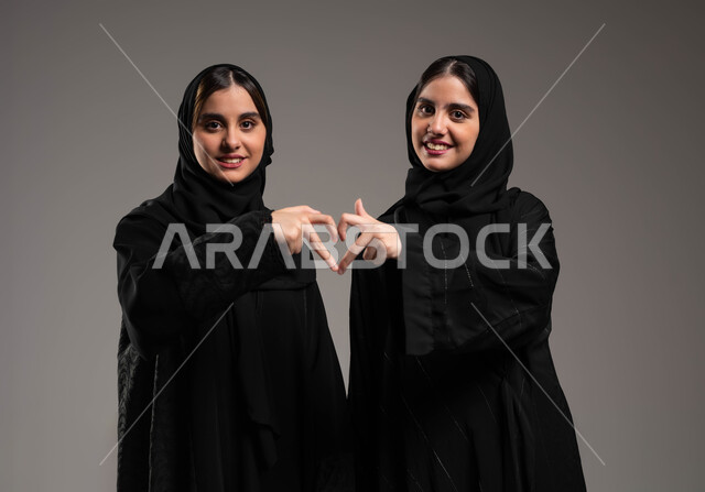 المحبة والمودة بين الشقيقات، بورتريه مقرب لاختين توءم عربيتين خليجيتين سعوديتين محجبتين مبتسمتين ترتديان العباءة السوداء وتشكلن قلب حب بيديهما تنظران الى الكاميرا بتعابير السرور، تقوية وتعزيز الروابط العائلية، معنى الصداقة والاخوة، خلفية رمادية