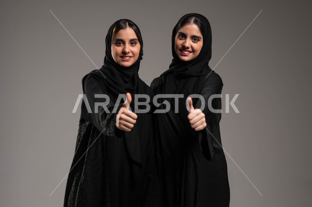 النظر إلى الكاميرا بتعابير السرور، القبول والرضا بالنتيجة، ايماءات الاعجاب والموافقة، بورتريه لأختين توءم عربيتين خليجيتين سعوديتين محجبتين مبتسمتين ترتديان العباءة ترفعان اصبع الابهام للأعلى بعلامة الجودة والامتياز، الاتفاق على أمر معين، خلفية رمادية