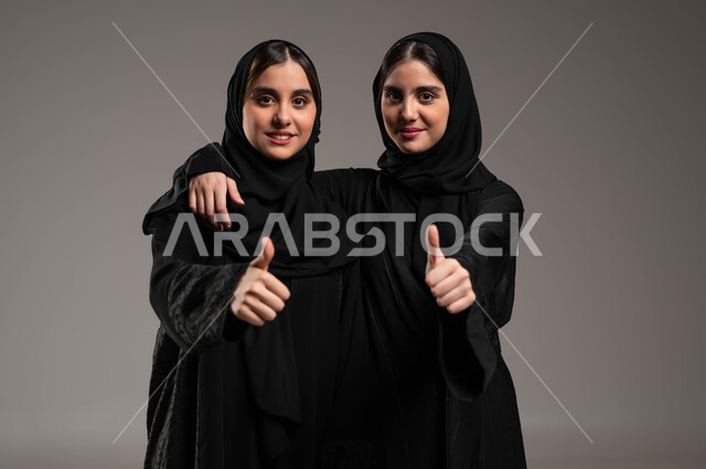شابة تعانق اختها، النظر إلى الكاميرا بتعابير السرور، القبول والرضا بالنتيجة، ايماءات الاعجاب والموافقة، بورتريه مقرب لأختين توءم عربيتين خليجيتين سعوديتين محجبتين ترتديان العباءة ترفعان اصبع الابهام للأعلى بعلامة الجودة، الاتفاق على أمر معين، خلفية رمادية