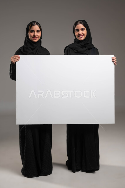 النظر إلى الكاميرا بإيماءات السرور والسعادة، استخدام لوحات توضيحية، بورتريه لتوءم بنات عربيات خليجيات سعوديات محجبات مبتسمات ترتديان العباءة تحملان لوح اعلاني أبيض فارغ، موك اب مستطيل كبير، صورة الجسم بالطول الكامل، خلفية رمادية