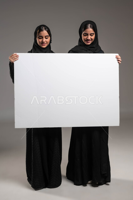 استخدام لوحات توضيحية، بورتريه لتوءم بنات عربيات خليجيات سعوديات محجبات مبتسمات ترتديان العباءة تحملان لوح اعلاني أبيض فارغ تنظران إليه بإيماءات البشاشة، موك اب مستطيل كبير، صورة الجسم بالطول الكامل، خلفية رمادية