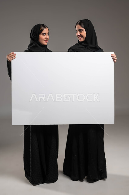 مفهوم التوءم الحقيقي المتطابق، استخدام لوحات توضيحية، بورتريه لتشابتين عربيتين خليجيتين سعوديتين مبتسمتين ترتديان العباءة تحملان لوح اعلاني أبيض فارغ تنظران لبعضهما بإيماءات البشاشة والسرور، موك اب مستطيل كبير، صورة الجسم بالطول الكامل، خلفية رمادية