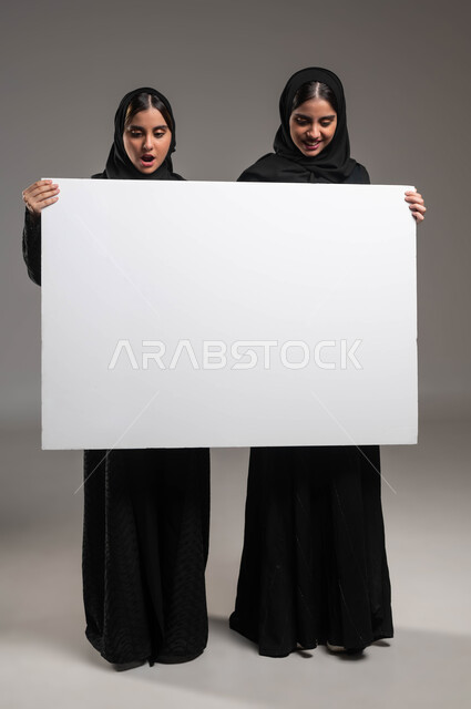 شابة سعودية تمسك بيدها لوح اعلاني أبيض فارغ تنظر إليه بإيماءات المفاجأة والتعجب، استخدام لوحات توضيحية، بورتريه لتوءم بنات عربيات خليجيات سعوديات محجبات مبتسمات ترتديان العباءة تحملان موك اب مستطيل كبير، صورة الجسم بالطول الكامل، خلفية رمادية