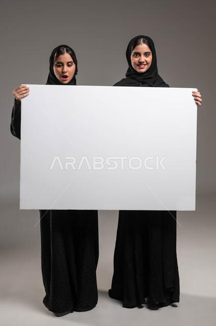 شابة سعودية تمسك بيدها لوح اعلاني أبيض فارغ تنظر إليه بإيماءات المفاجأة والتعجب، استخدام لوحات توضيحية، بورتريه لتوءم بنات عربيات خليجيات سعوديات محجبات مبتسمات ترتديان العباءة تحملان موك اب مستطيل كبير، صورة الجسم بالطول الكامل، خلفية رمادية