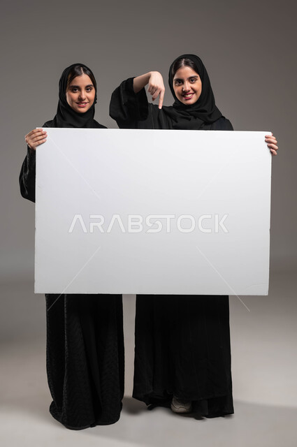 النظر إلى الكاميرا بإيماءات السرور، الإشارة بإصبع السبابة إلى لوح اعلاني أبيض فارغ، استخدام لوحات توضيحية، بورتريه لتوءم بنات عربيات خليجيات سعوديات محجبات مبتسمات ترتديان العباءة تحملان موك اب مستطيل كبير، صورة الجسم بالطول الكامل، خلفية رمادية
