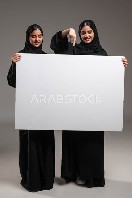 الإشارة بإصبع السبابة إلى لوح اعلاني أبيض فارغ، استخدام لوحات توضيحية، بورتريه لتوءم بنات عربيات خليجيات سعوديات محجبات ترتديان العباءة تحملان موك اب مستطيل كبيرتنظران إليه باهتمام، صورة الجسم بالطول الكامل، خلفية رمادية