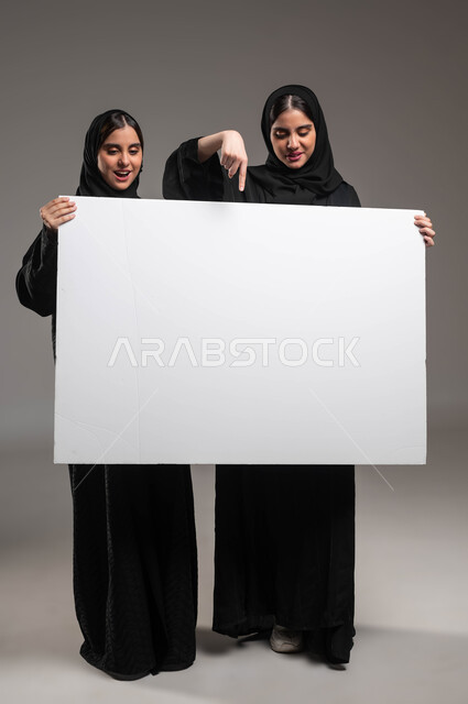 لوح اعلاني فارغ، الإشارة بإصبع السبابة بإيماءات التعجب والمفاجأة، استخدام لوحات توضيحية ووسائل تعليمية، إعلانات وعروض تسويقية، بورتريه لتوءم بنات عربيات خليجيات سعوديات محجبات ترتديان العباءة تحملان موك اب مستطيل كبير، صورة الجسم بالطول الكامل، خلفية رمادية