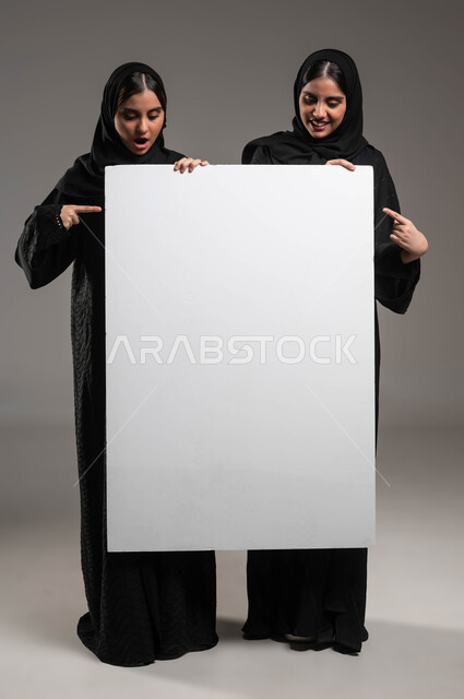 الإشارة بإصبع السبابة بإيماءات التعجب والمفاجأة، لوح اعلاني فارغ، استخدام لوحات توضيحية ووسائل تعليمية، إعلانات وعروض تسويقية، بورتريه لتوءم بنات عربيات خليجيات سعوديات محجبات ترتديان العباءة تحملان موك اب مستطيل كبير، صورة الجسم بالطول الكامل، خلفية رمادية