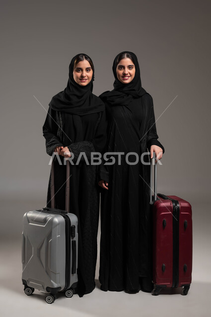 السياحة في إجازة الصيف, الاستعداد لقضاء رحلة صيفية ممتعة, بورتريه لشابتين توءم عربيتين خليجيتين سعوديتين مبتسمتين محجبتين ترتديان العباءة السوداء تمسكات حقيبة السفر وتنظران إلى الكاميرا بإيماءات السرور، صورة لكامل الجسم, خلفية رمادية