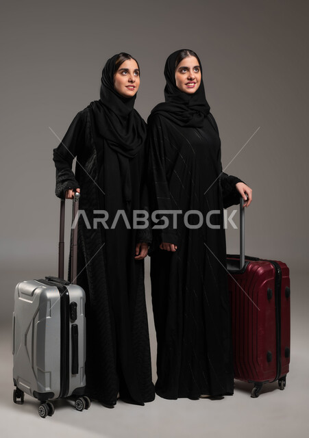 الاستعداد لقضاء رحلة صيفية ممتعة, السياحة في إجازة الصيف, بورتريه لشابتين توءم عربيتين خليجيتين سعوديتين مبتسمتين محجبتين ترتديان العباءة السوداء تمسكات حقيبة السفر وتنظران للأعلى بإيماءات السرور، صورة لكامل الجسم, خلفية رمادية