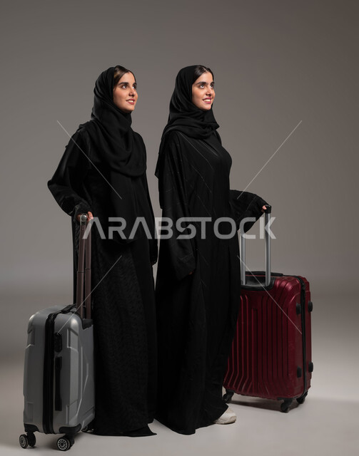 السياحة في إجازة الصيف, الاستعداد لقضاء رحلة صيفية ممتعة, بورتريه لشابتين توءم عربيتين خليجيتين سعوديتين مبتسمتين محجبتين ترتديان العباءة السوداء تمسكات حقيبة السفر وتنظران للأعلى بإيماءات السرور، صورة لكامل الجسم, خلفية رمادية