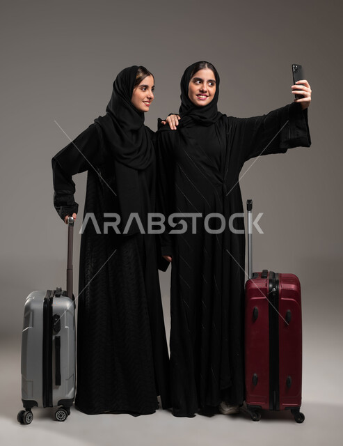 الاستعداد لقضاء رحلة صيفية ممتعة، السياحة في إجازة الصيف, بورتريه لشابة عربية خليجية سعودية محجبة ترتدي العباءة تقوم بالتقاط صور السيلفي التذكارية مع اختها التوءم باستخدام الجوال بإيماءات السرور، صورة الجسم بالكامل، خلفية رمادية