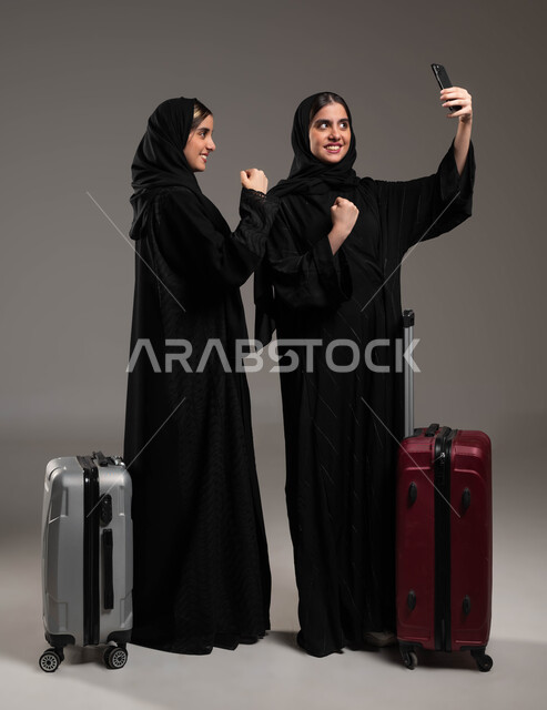 السياحة في إجازة الصيف, الاستعداد لقضاء رحلة صيفية ممتعة، بورتريه لشابة عربية خليجية سعودية محجبة ترتدي العباءة السوداء تقوم بالتقاط صور السيلفي التذكارية مع اختها التوءم باستخدام الجوال بإيماءات السرور والفوز، صورة الجسم بالكامل، خلفية رمادية