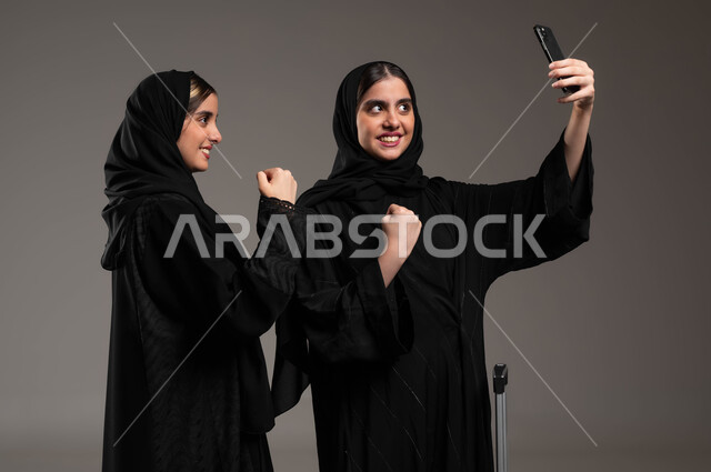 الإشارة بعلامة الفوز والانتصار، توثيق اللحظات السعيدة، استخدام الأجهزة التقنية المتطورة، بورتريه مقرب لشابة عربية خليجية سعودية محجبة ترتدي العباءة السوداء تقوم بالتقاط صور السيلفي التذكارية مع اختها التوءم بإيماءات السعادة والسرور، خلفية رمادية