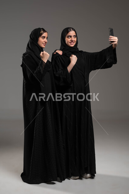 توثيق اللحظات السعيدة، الإشارة بعلامة الفوز والانتصار، استخدام الأجهزة التقنية المتطورة، بورتريه لشابة عربية خليجية سعودية محجبة ترتدي العباءة السوداء تقوم بالتقاط صور السيلفي التذكارية مع اختها التوءم بإيماءات السعادة والسرور، صورة الجسم بالكامل، خلفية رمادية