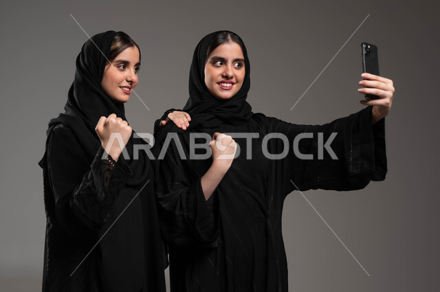 الإشارة بعلامة الفوز والانتصار، توثيق اللحظات السعيدة، استخدام الأجهزة التقنية المتطورة، بورتريه مقرب لشابة عربية خليجية سعودية محجبة ترتدي العباءة السوداء تقوم بالتقاط صور السيلفي التذكارية مع اختها التوءم بإيماءات السعادة والسرور، خلفية رمادية
