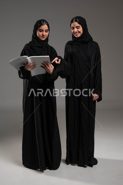 ثقافة التعلم الذاتي، مراجعة الدروس اليومية، الاستمتاع بممارسة هواية المطالعة، بورتريه لبنتين توءم عربتين خليجيتين سعوديتين محجبتين مبتسمتين تقضيان وقتهما بالقراءة والتثقف، شابة تشير بإصبع السبابة إلى الكتاب، صورة الجسم بالكامل، خلفية رمادية